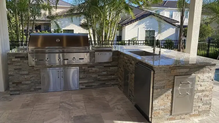 Outdoor kitchen with a built in grill, stone counters and a stainless sink under a covered lanai